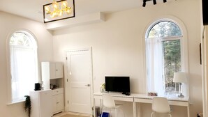 A room with a desk, two chairs, a television, a white wardrobe, a coffee maker, and a fire extinguisher.