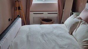 Et soveværelse med seng, gulvlampe, et vindue med gardiner, en radiator og en lille kurvefletkurv.