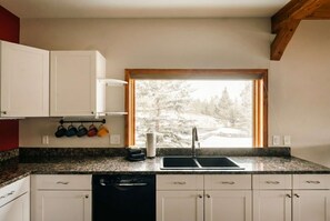 Une cuisine avec des meubles blancs, un plan de travail en granit, une fenêtre donnant sur des arbres couverts de neige et un égouttoir à vaisselle rempli de tasses colorées.