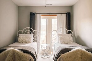 Quarto com duas camas de metal branco, uma mesa redonda pequena e vista para o exterior através de uma janela com cortinas.