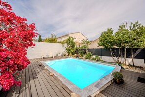 A swimming pool with a wooden deck, surrounded by plants and a house in the background. - Ground Floor of a Villa in Toulon - Private Pool (Toulon)