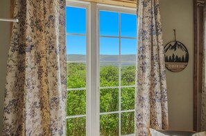 Ein Zimmer mit einem großen Fenster, blumigen Vorhängen, Blick ins Grüne und einer Wanddekoration mit Bergen und Bäumen.