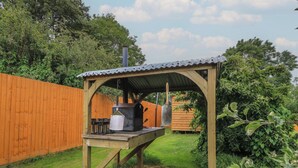 A wooden outdoor barbecue setup with a metal roof, a grill, and a pot hanging from the structure. - Damsons (Taunton)