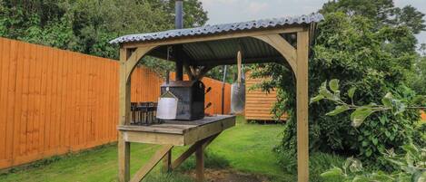 Una barbacoa exterior de madera con techo metálico, una parrilla y una olla colgando de la estructura.