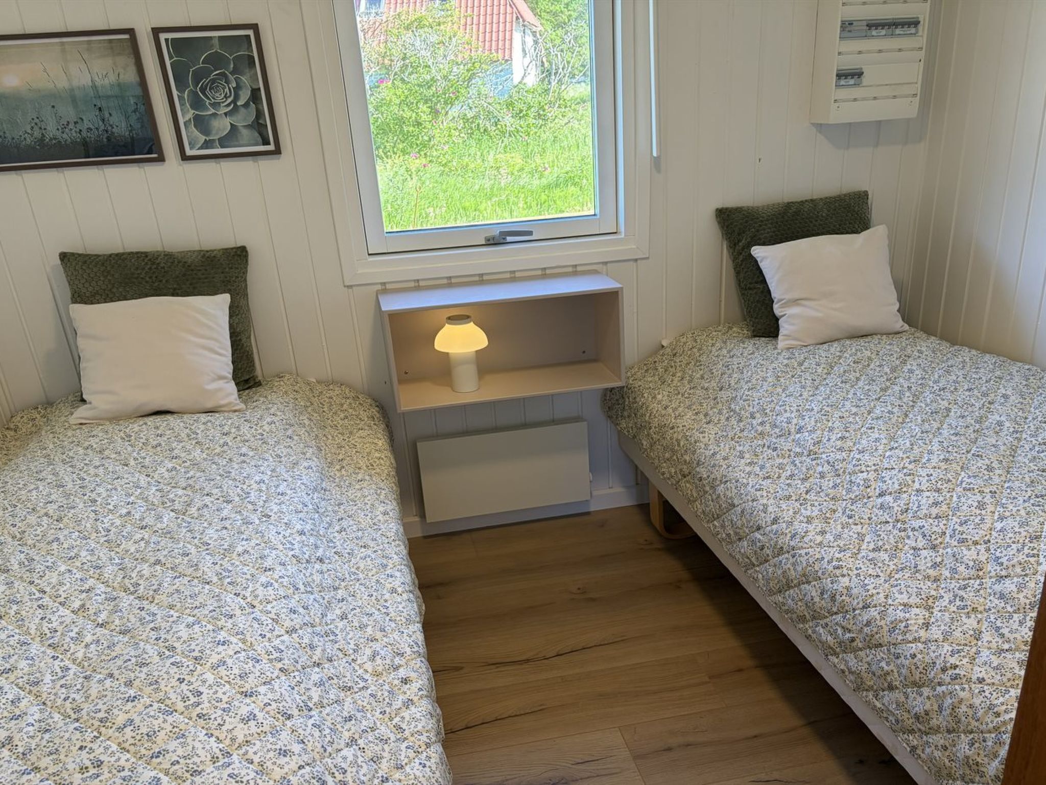 Een kamer met twee éénpersoonsbedden, een nachtkastje met een lamp, een raam met uitzicht op groen, en een wandpaneel met elektrische installatie.