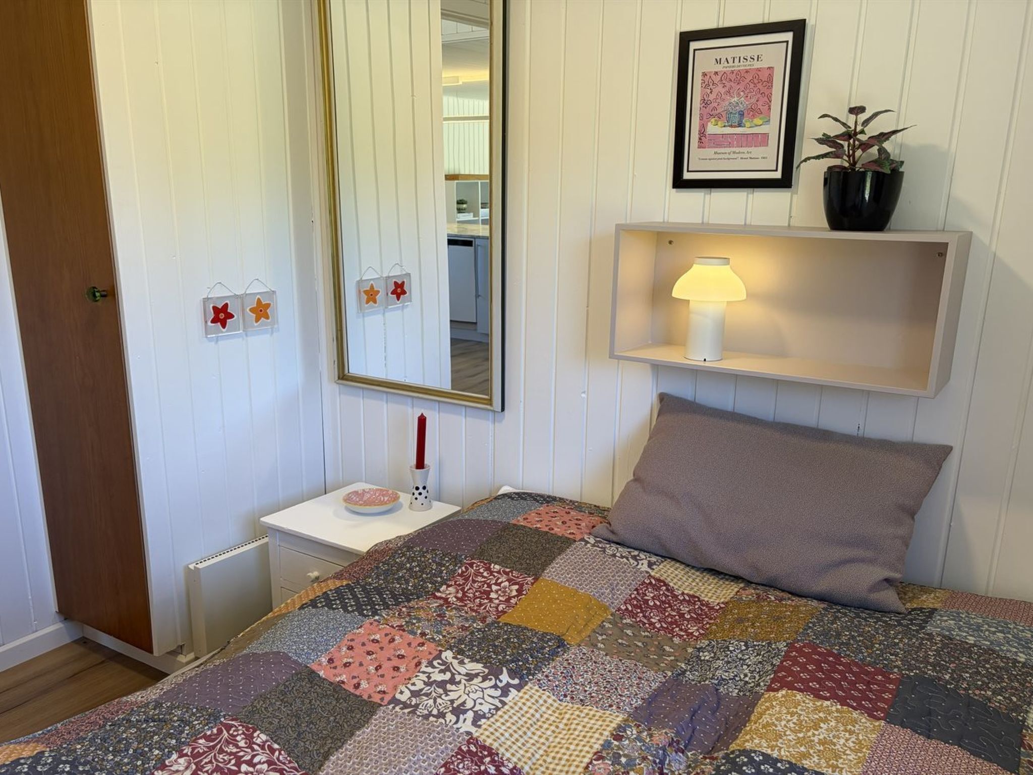 A bedroom with a quilted bedspread, a bedside lamp, a mirror, and a framed artwork on the wall.