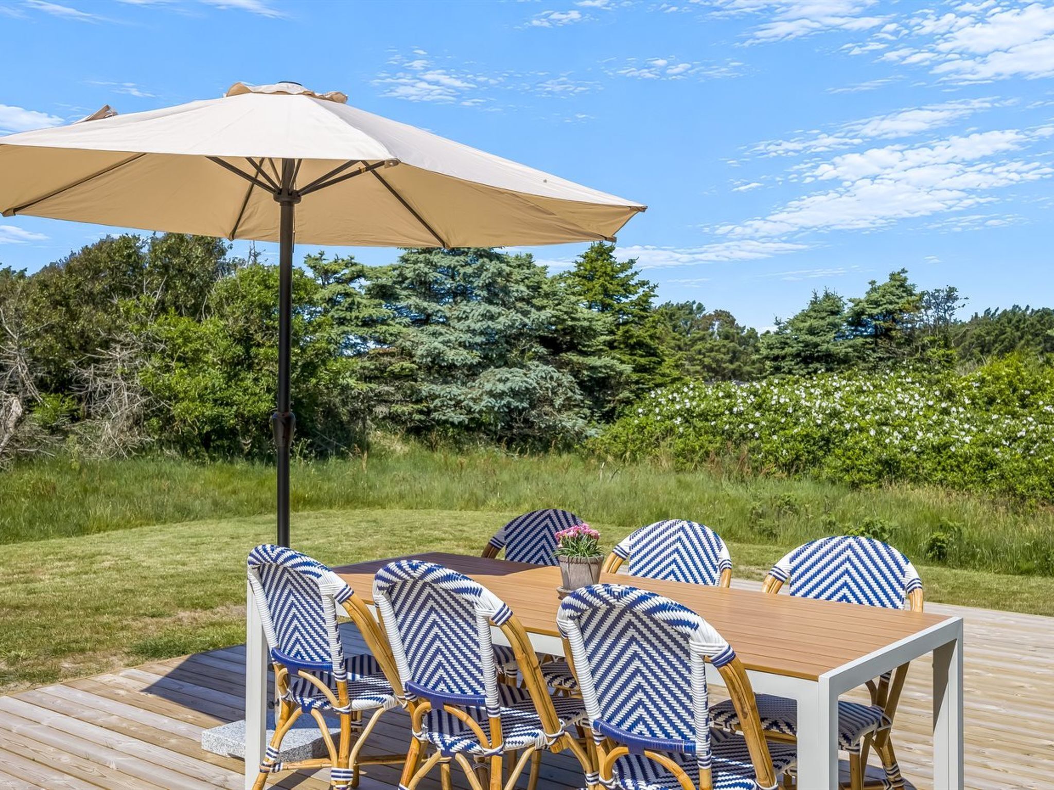 An outdoor dining area with a wooden deck, a table, six chairs with patterned cushions, and a large beige umbrella.