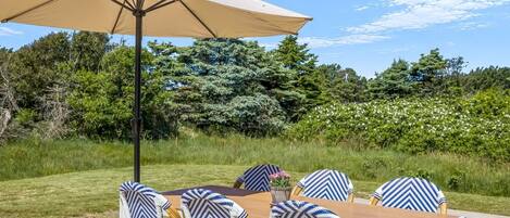 An outdoor dining area with a wooden deck, a table, six chairs with patterned cushions, and a large beige umbrella.