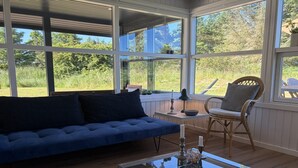Una habitación con sofá azul, una mesa de centro de vidrio, sillas de mimbre y ventanas amplias con vistas a zonas verdes.