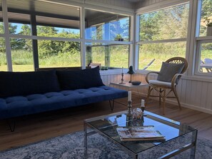 Ein Zimmer mit einem blauen Sofa, einem Glastisch, Korbstühlen und großen Fenstern mit Blick ins Grüne.