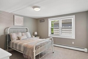 A bedroom with a metal bed frame, a bedside table with a lamp, a window with blinds, and a wall-mounted sign. - Walloon Waypoint - Walloon Lake (Bear Creek Township)