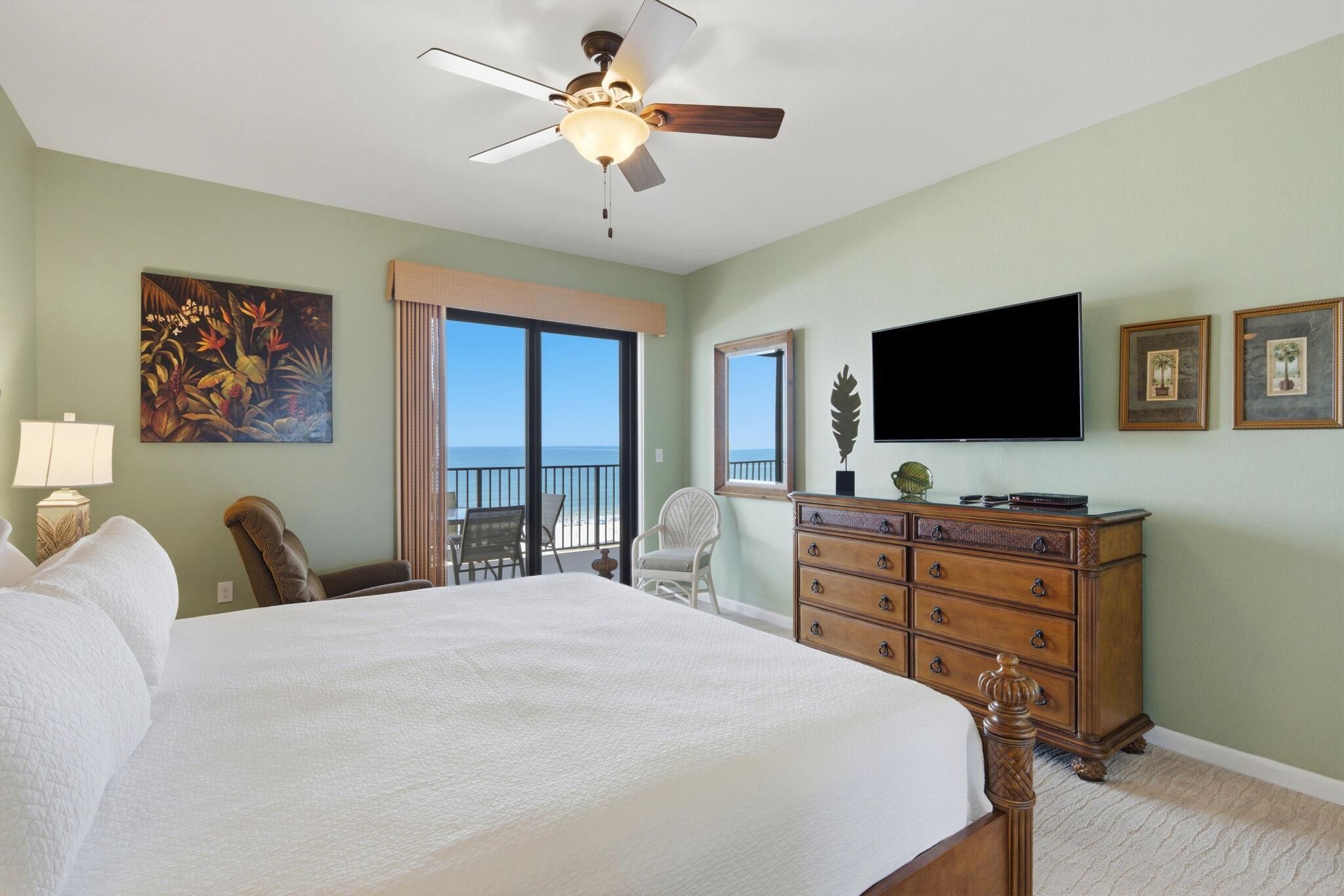 A hotel room with a large bed, a ceiling fan, a TV, a dresser, a chair, and a balcony with a view.