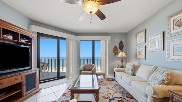 A living room with a television, a ceiling fan, a sofa, a coffee table, and a view of the ocean through large windows. - Phoenix 6 1111 (Orange Beach)