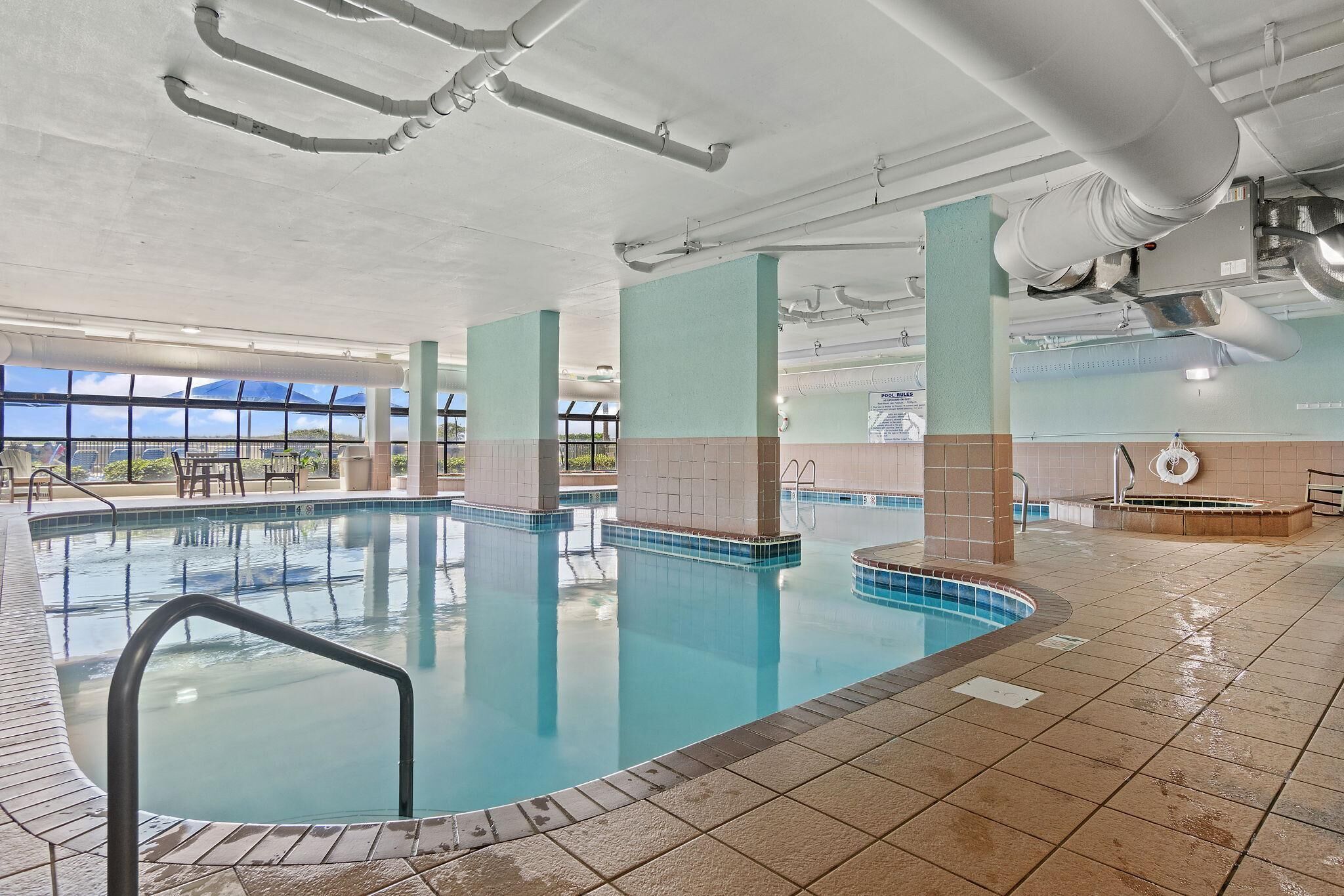 An indoor swimming pool with clear blue water, tiled walls, and a view of the sky through large windows.