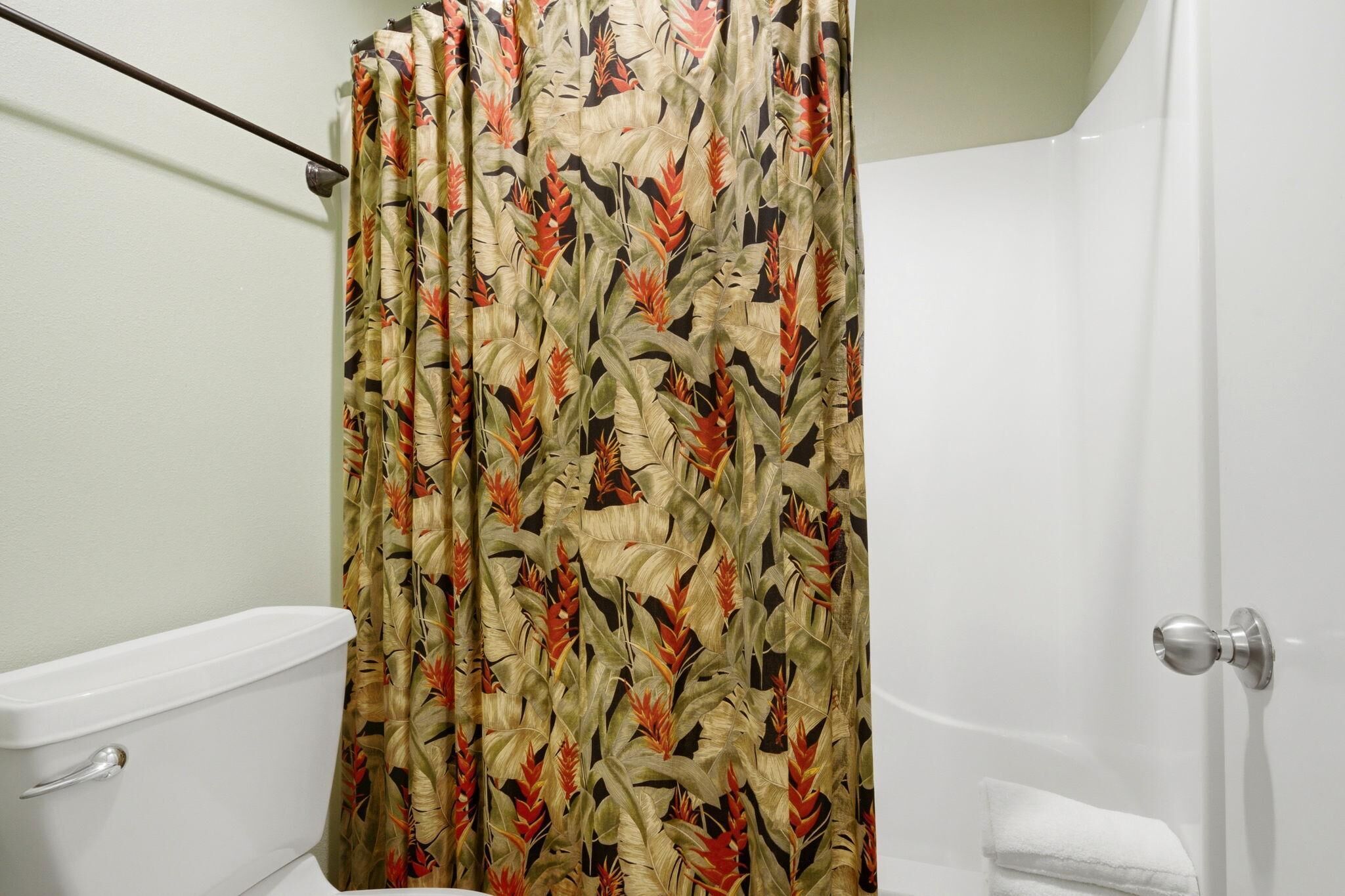 A bathroom with a toilet, a shower curtain with a tropical leaf pattern, and a white towel on the floor.