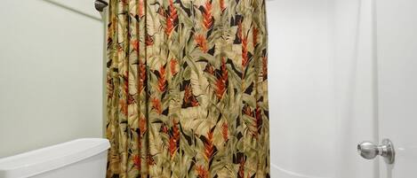A bathroom with a toilet, a shower curtain with a tropical leaf pattern, and a white towel on the floor.