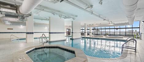An indoor swimming pool with a hot tub, clear ceiling with ventilation ducts, and large windows providing natural light.