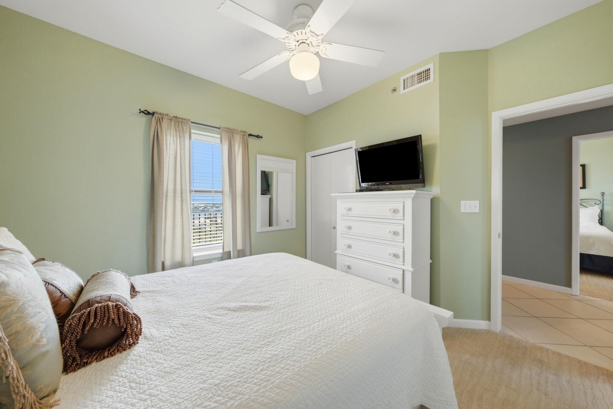 A bedroom with a bed, a TV, a ceiling fan, and a window with curtains.