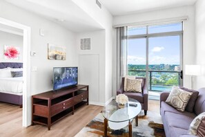 Ein modernes Wohnzimmer mit Fernseher, Sofa und Ohrensessel, einem Glastisch und einem großen Fenster mit Blick auf die Stadt.