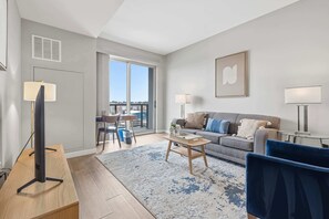 Ein modernes Wohnzimmer mit Sofa, Couchtisch und Fernseher. Ein Balkon bietet Blick auf Gebäude.