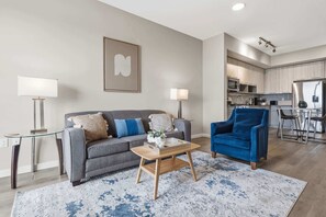 Een moderne woonkamer met een grijze bank, een blauwe fauteuil, een salontafel en een keuken op de achtergrond.