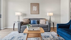 Ein modernes Wohnzimmer mit einem grauen Sofa, zwei Beistelltischen mit Lampen, einem blauen Sessel und einem Couchtisch mit einem Blumenstrauß.