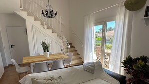 Un salon moderne avec un canapé, une table à manger en bois, des chaises blanches, un escalier et une vue à travers une porte vitrée.