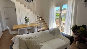 Una sala de estar moderna con un sofá, una mesa de comedor de madera, sillas blancas, una escalera y vistas a través de un ventanal.