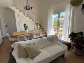 Ein modernes Wohnzimmer mit einer Couch, einem Holzesstisch, weißen Stühlen, einer Treppe und Blick durch eine Glastür.