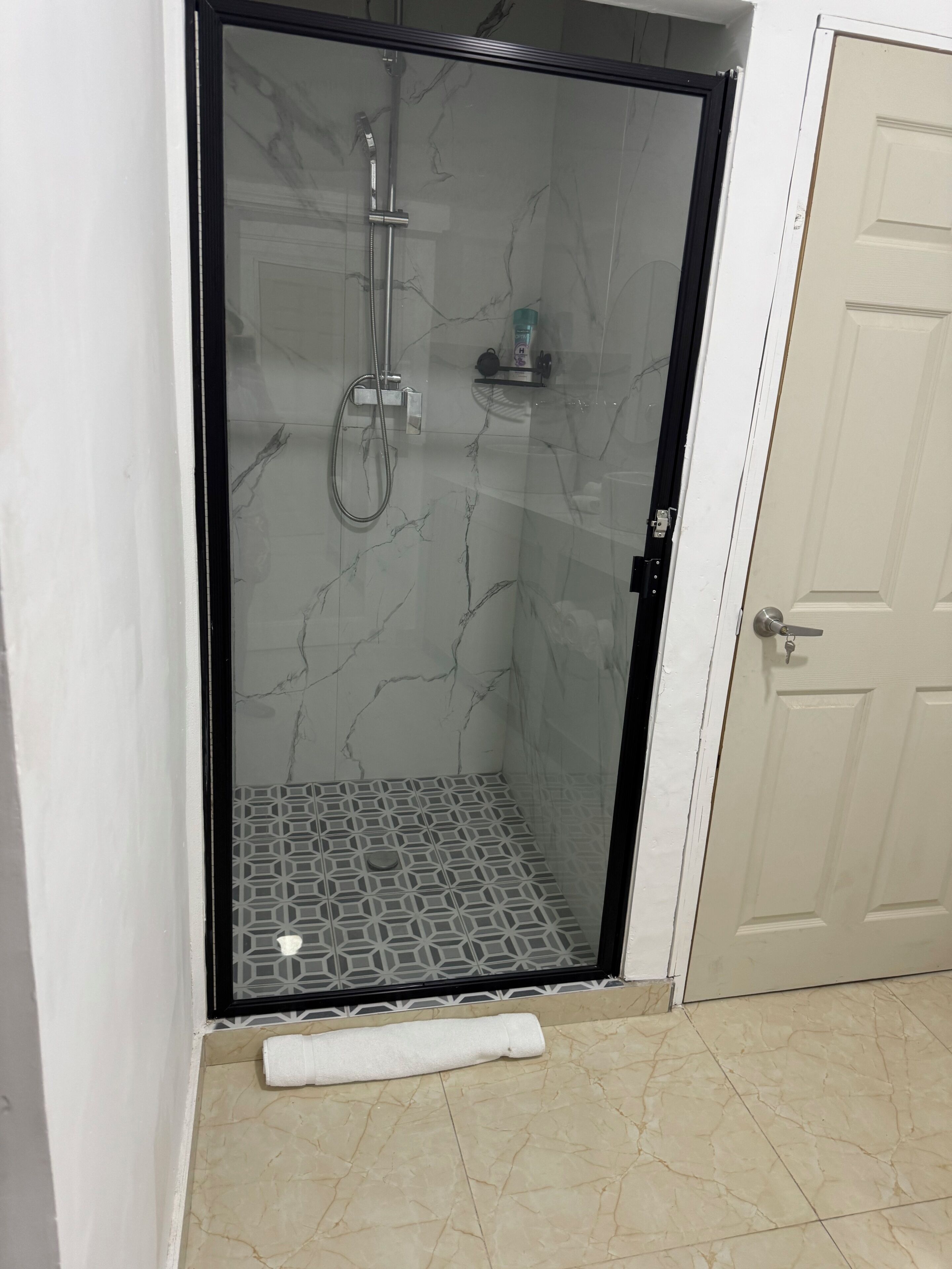 Una cabina doccia in vetro con cornice nera, pavimento piastrellato e una porta bianca adiacente.
