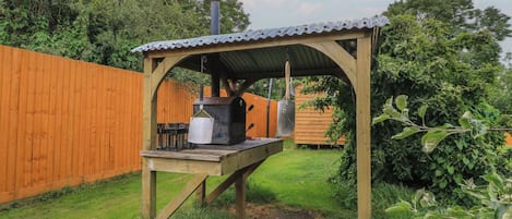 A wooden outdoor barbecue setup with a metal roof, a grill, and a pot hanging from the structure.