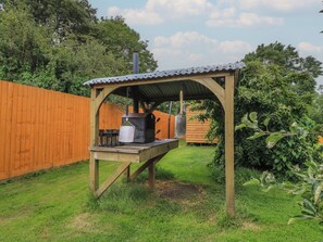 Eine hölzerne Grillanlage im Freien mit Metallüberdachung, einem Grill und einem daran hängenden Topf.