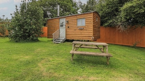 A small wooden cabin with a door and windows, a staircase, and a picnic table in a grassy yard. - Pippins (Taunton)