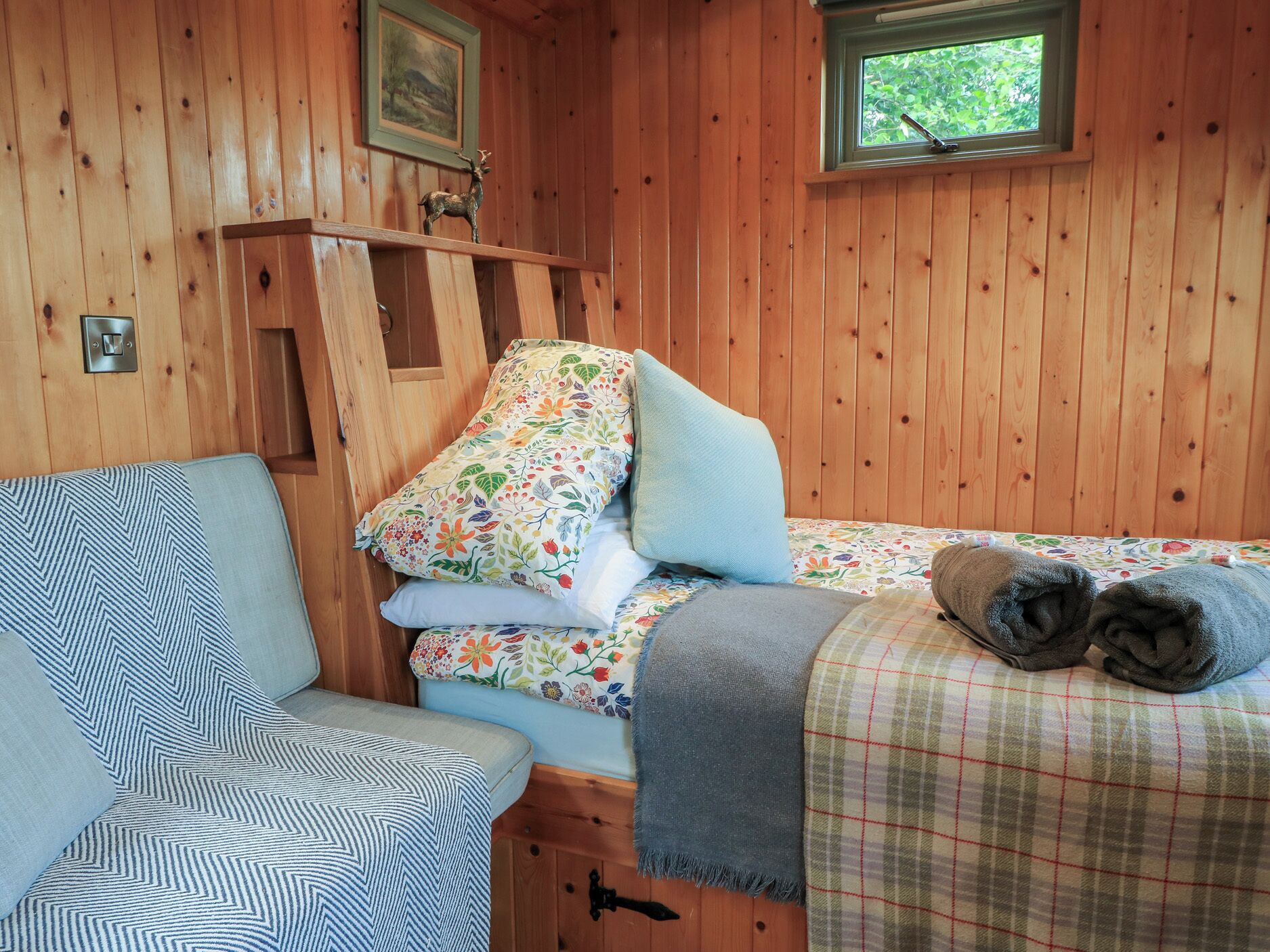 Interior de una cabaña de madera con una cama, una silla, una ventana y una repisa con una figurita de venado.