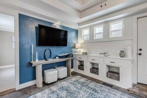 Una sala de estar moderna con un televisor de pantalla plana grande montado en una pared azul, una consola blanca, dos zonas de descanso con cojines blancos y una alfombra estampada.