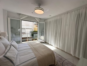 Een slaapkamer met een bed, een plafondventilator, een raam met gordijnen en uitzicht op buiten.