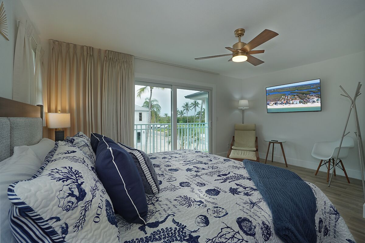A bedroom with a bed, curtains, a ceiling fan, a window with a view, a chair, and a small table.