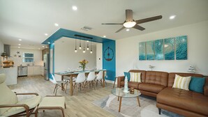 Un salon moderne avec un canapé en cuir marron, un coin repas avec des chaises blanches et une cuisine en arrière-plan.