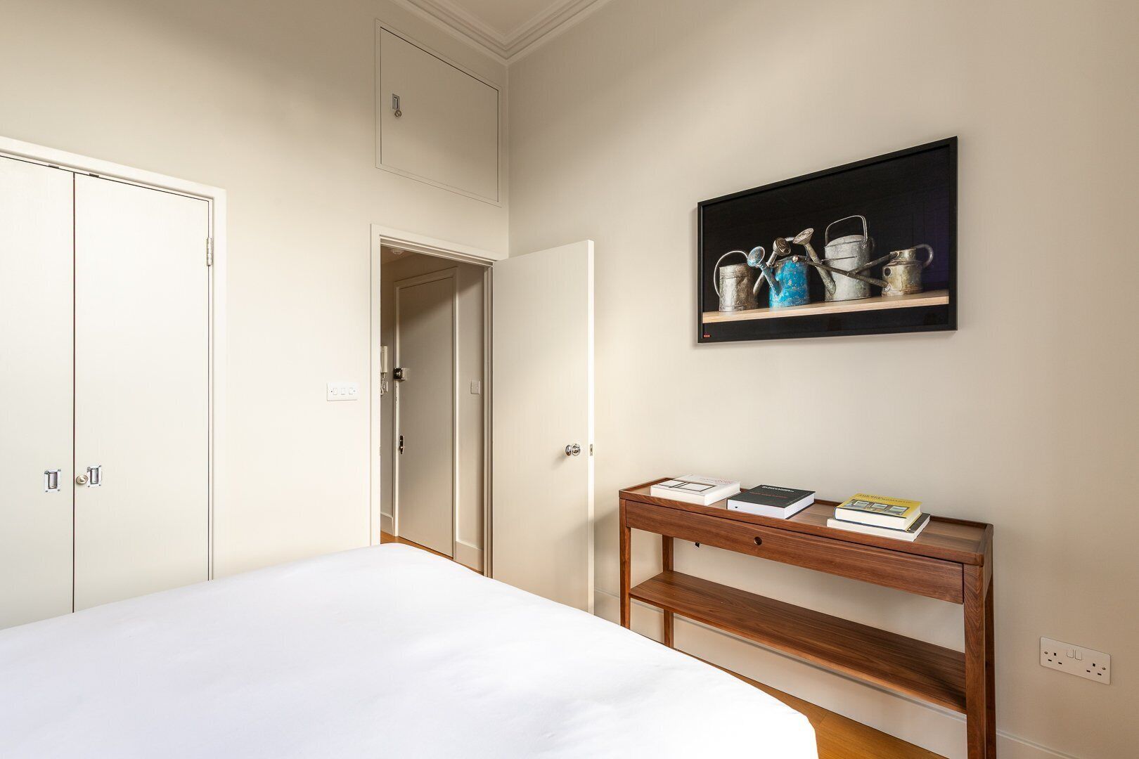 Un dormitorio con cama, una mesita de noche de madera con libros, un cuadro y un armario.