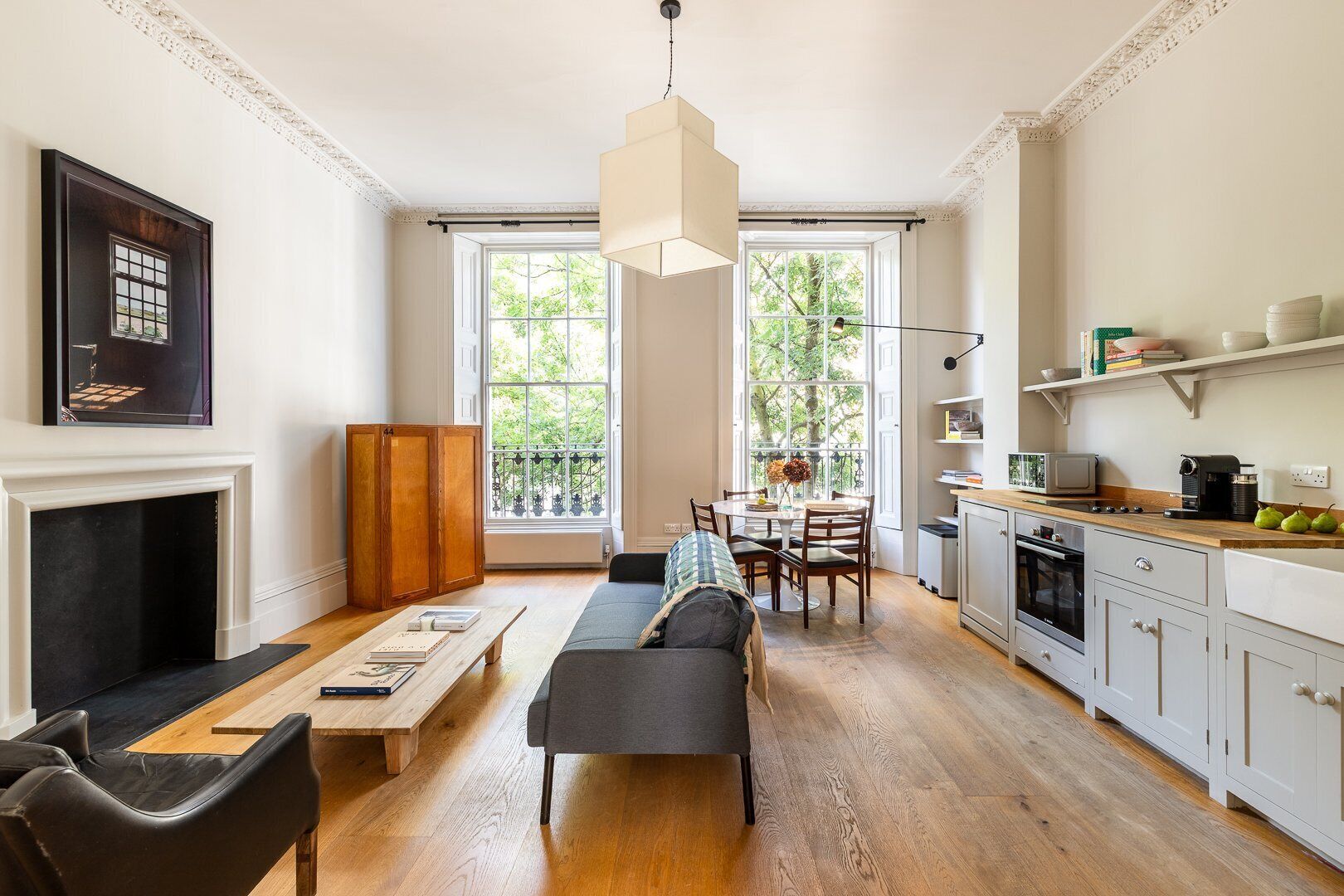 A spacious living room with a fireplace, a dining area with a table and chairs, a kitchen with a stove and cabinets, and large windows with a view of greenery.
