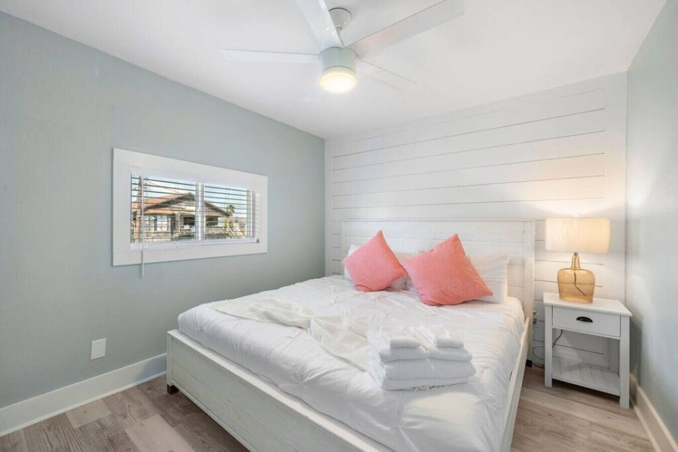 Um quarto com cama, mesa de cabeceira com abajur, janela com persianas e ventilador de teto.