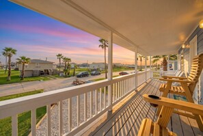 Una terraza de madera con sillas, vista a una calle con casas y un cielo al atardecer.