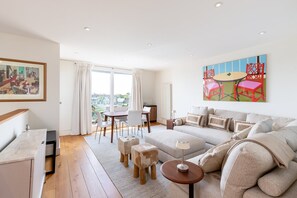 A modern living room with a sofa, a dining table, and chairs. There is a painting on the wall and a view of the outside. - Brook Green Flat by Kensington Olympia (London)