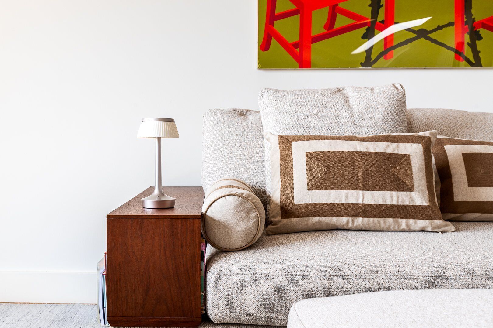 A neatly arranged bedroom with a wooden nightstand, a lamp, a sofa with patterned cushions, and a large abstract painting on the wall.