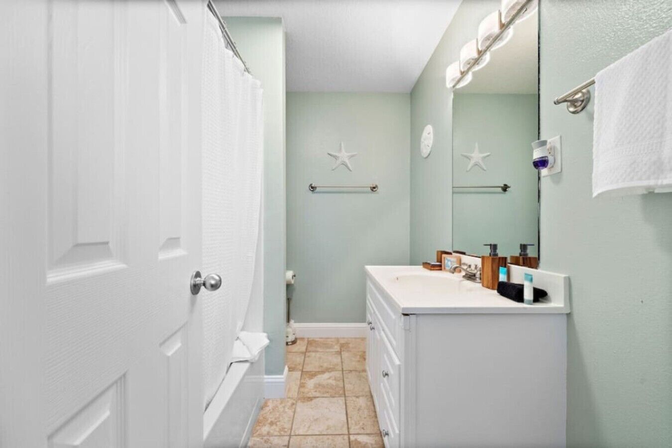 A bathroom with a white door, a sink with toiletries, a towel rack, and a mirror.