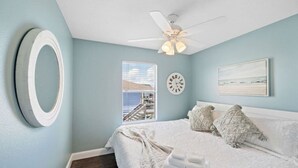A bedroom with a bed, a ceiling fan, a clock, a window with a view, and a painting on the wall.