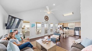 A spacious living room with a flat-screen TV, a ceiling fan, and a dining area with a kitchen in the background.