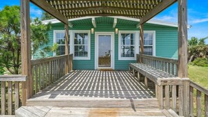Een houten terras leidt naar een huis met een groene gevel en een overdekte veranda.
