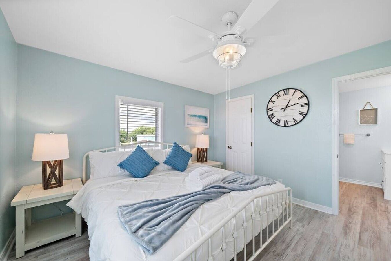 A bedroom with a bed, bedside tables, a ceiling fan, a wall clock, and a window with blinds.