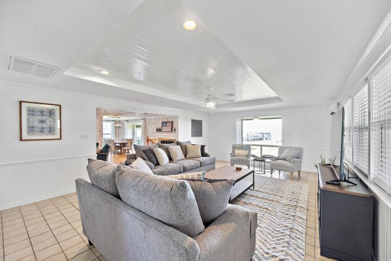 A spacious living room with a large grey sofa, a coffee table, and a TV stand. There is a dining area in the background with a table and chairs.